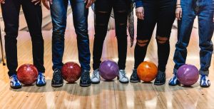 Five people are shown standing from the waist down with bowling balls between their feet.