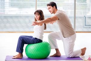 A girl sitting on a medicine ball next to her physical therapist with her arms pointing out.