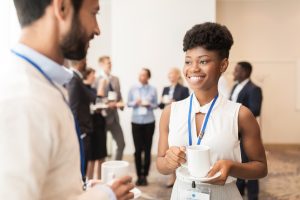 A woman is smiling, holding a cup of coffee, and is talking to a man. They are both at a conference